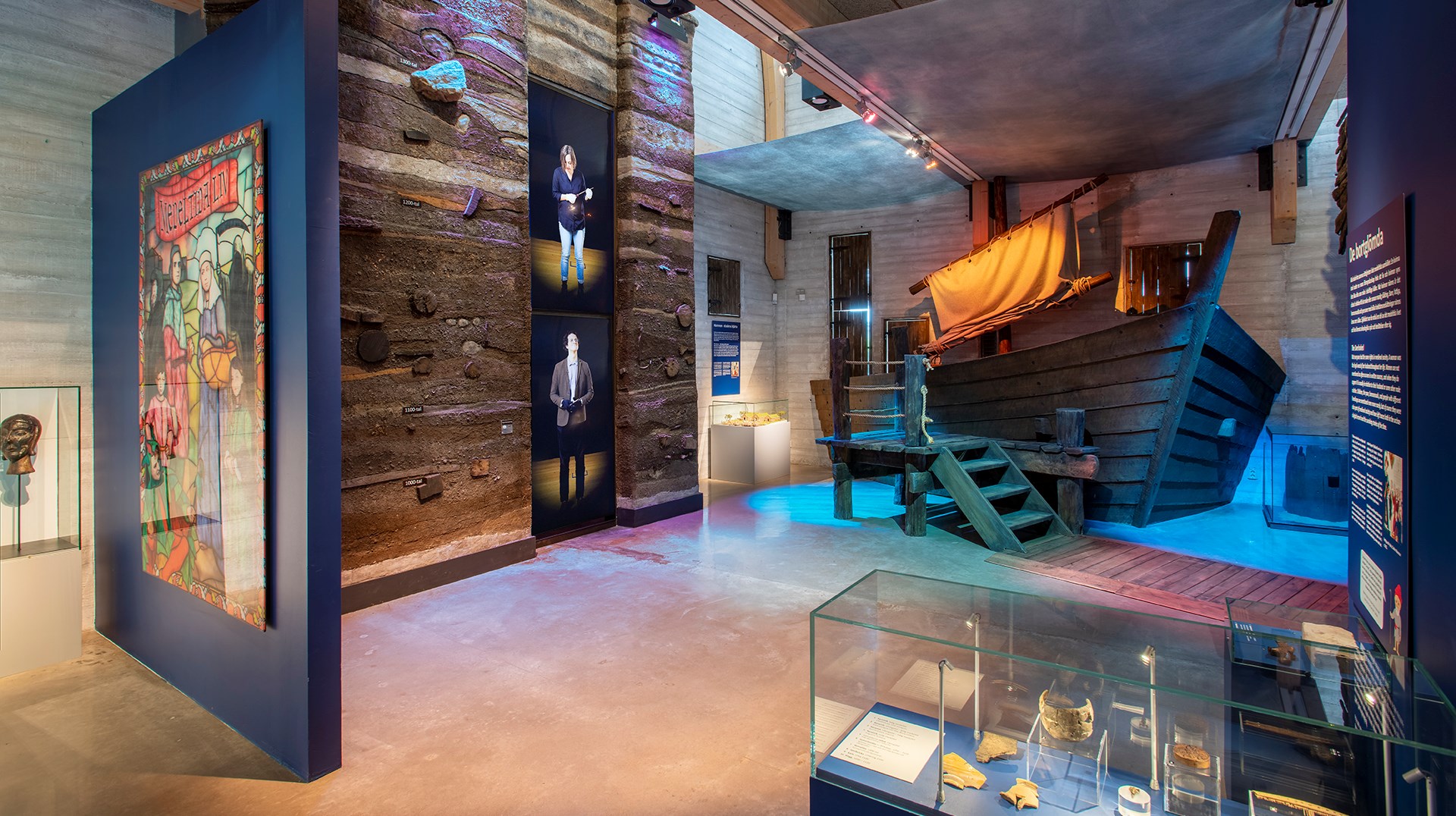 Översiktsbild över ett rum som kallas "ankomsten". Det är blå väggar, och en monter med föremål, en modell av en båt, och två stora skärmar där filmer spelas upp.