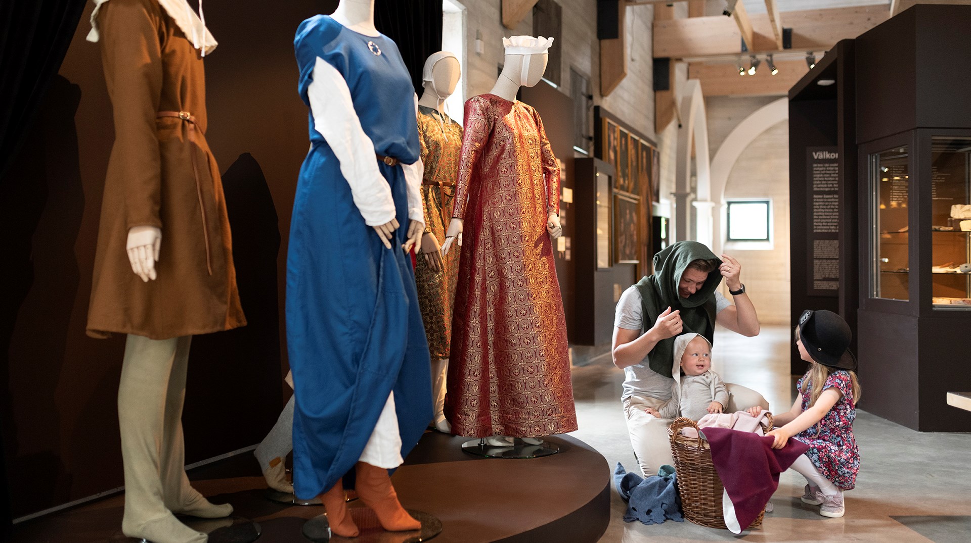 En man med två små barn sitter vid en korg och provar medeltida huvudbonader. Bredvid syns skyltdockor med medeltidskläder på sig