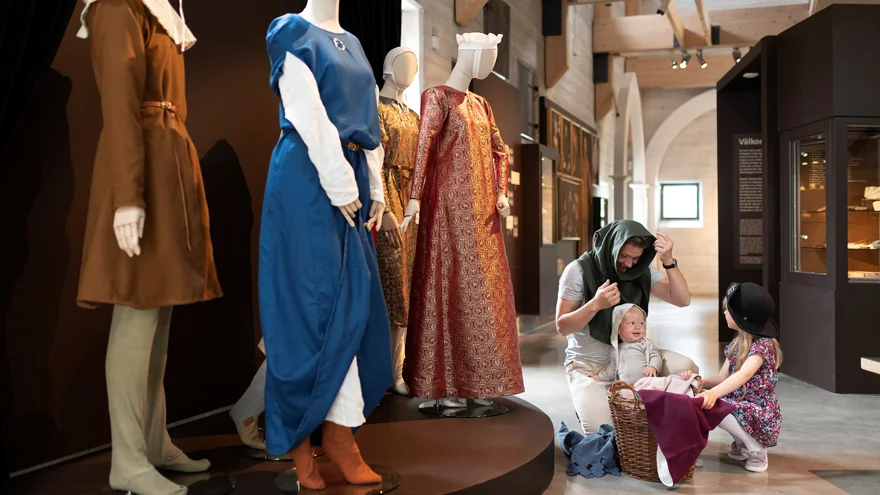 En man med två små barn sitter vid en korg och provar medeltida huvudbonader. Bredvid syns skyltdockor med medeltidskläder på sig