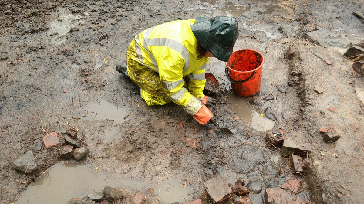 En arkeolog i regnkläder gräver i lerig jord