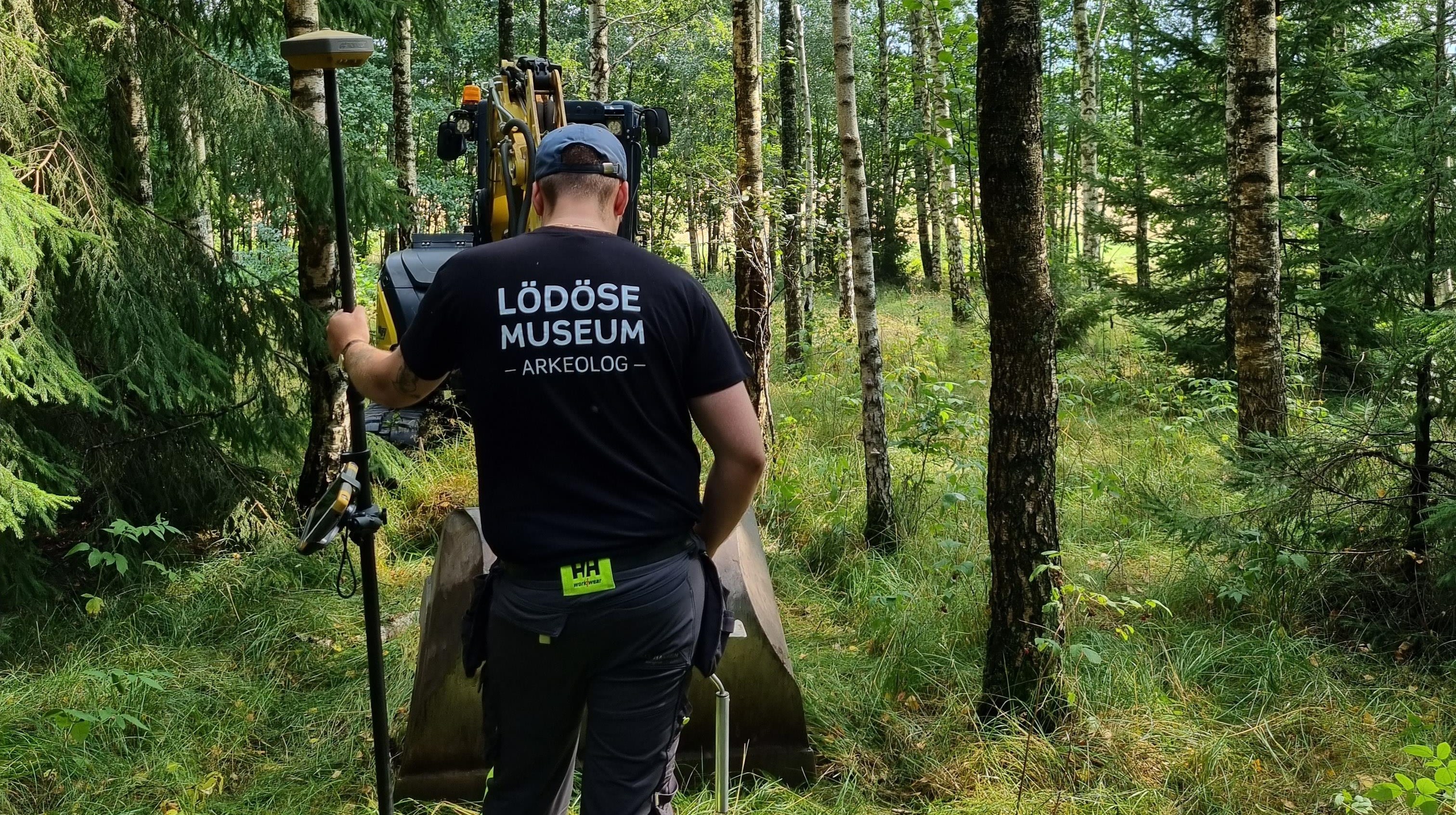 En arkeolog står i skogen och tittar på en grävmaskin som schaktgräver