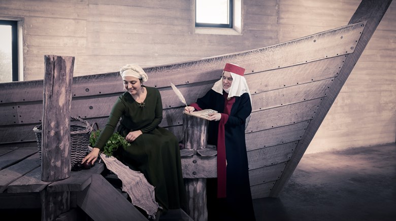 Två kvinnor är klädda i medeltidskläder och är framför en båt inomhus