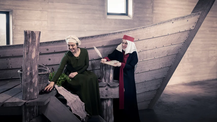 Två kvinnor är klädda i medeltidskläder och är framför en båt inomhus