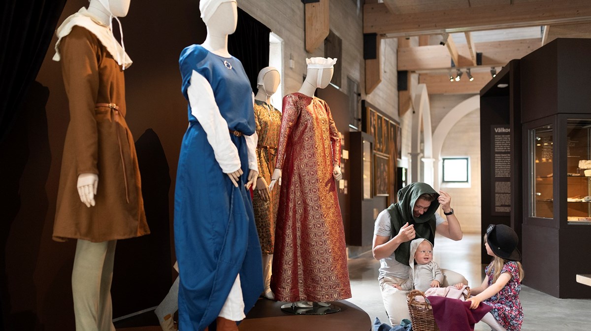 I förgrunden syns skyltdockor med medeltida kläder. Vid sidan av dem finns en man med två små barn som provar medeltida huvudbonader