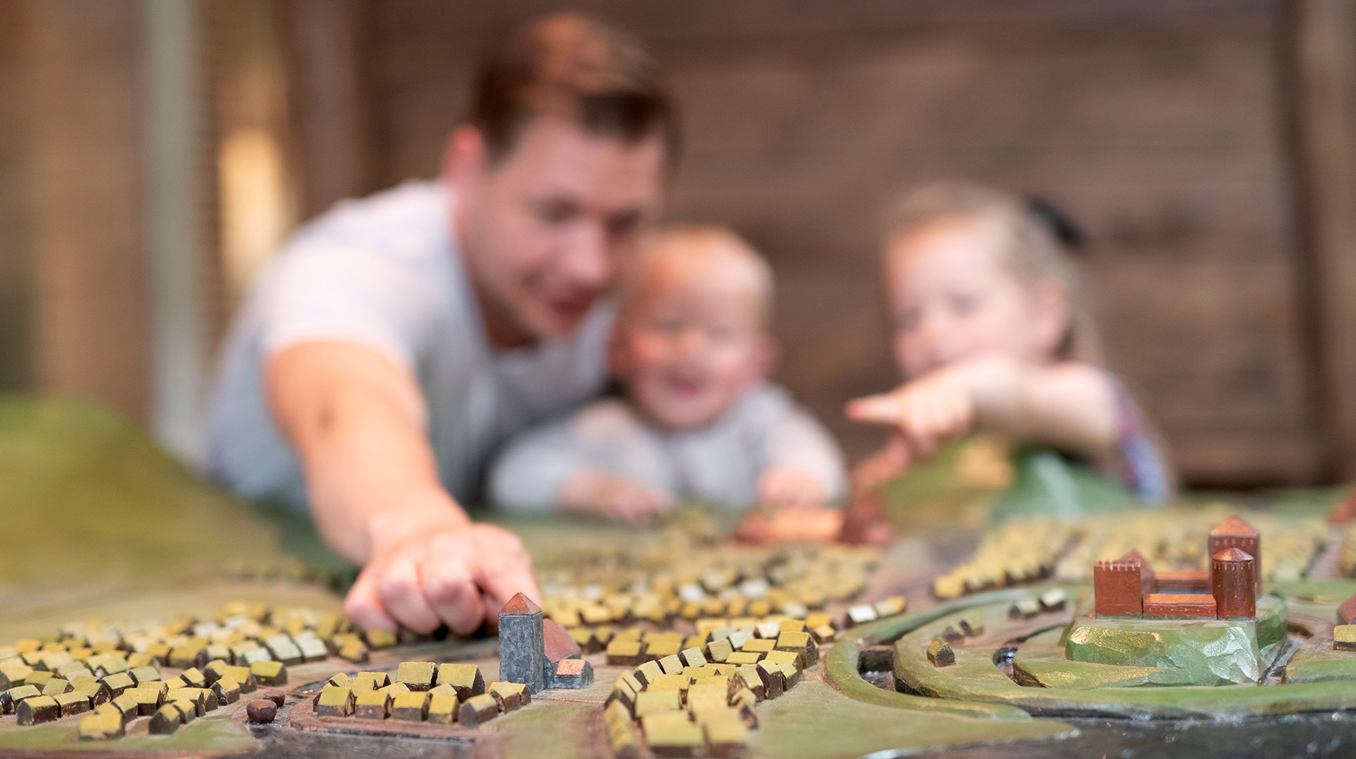 På bilden syns en pappa med sina två små barn suddigt i bakgrunden. I förgrunden ser man en trämodell över hus och kyrkor som ska föreställa Lödöse under medeltiden.