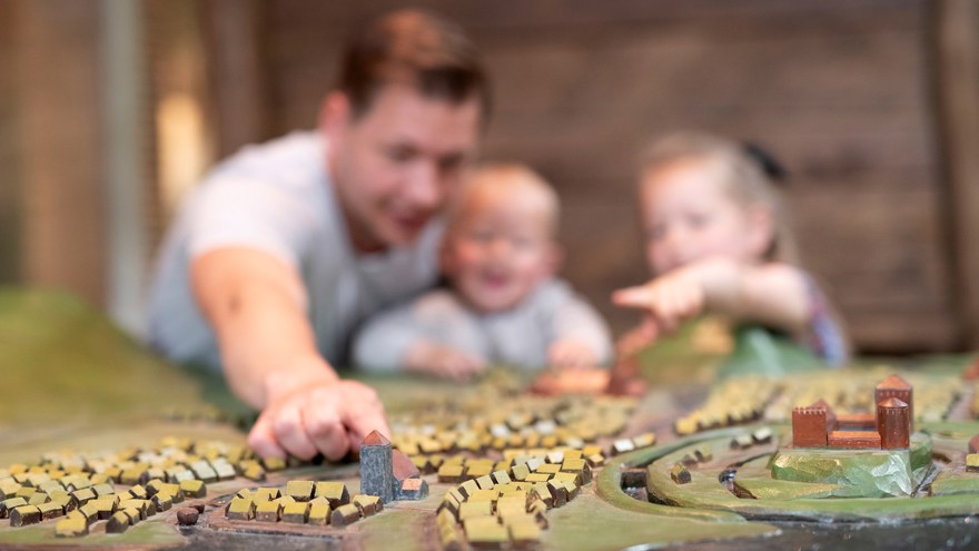 På bilden syns en pappa med sina två små barn suddigt i bakgrunden. I förgrunden ser man en trämodell över hus och kyrkor som ska föreställa Lödöse under medeltiden.