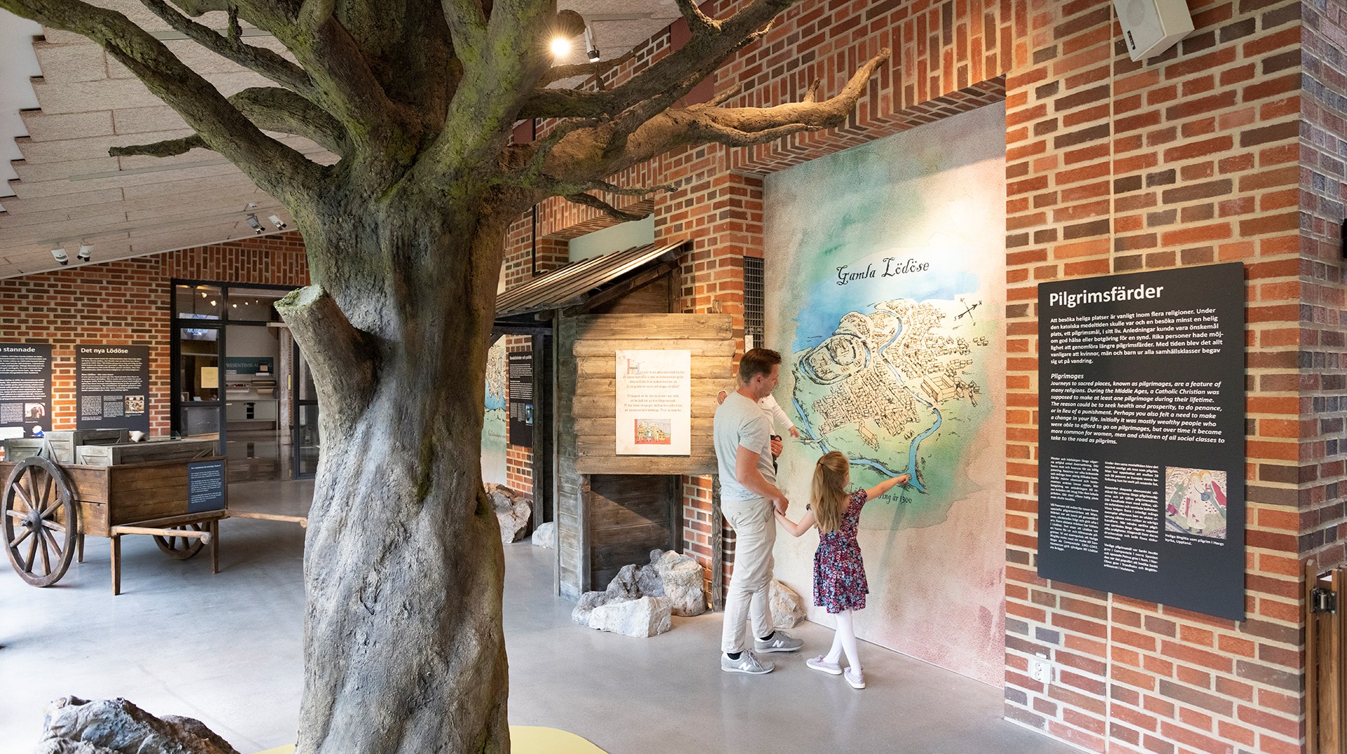 På bilden syns en pappa med sitt barn. De står i ett stort ljust rum med tegelväggar. I förgrunden syns ett träd. I bakgrunden står en kärra av trä. Pappan och barnet tittar på en karta som sitter på väggen.