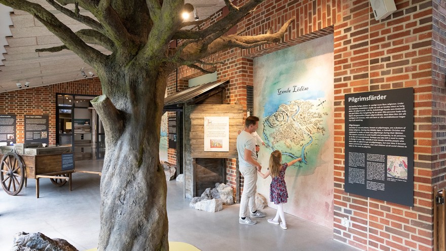 På bilden syns en pappa med sitt barn. De står i ett stort ljust rum med tegelväggar. I förgrunden syns ett träd. I bakgrunden står en kärra av trä. Pappan och barnet tittar på en karta som sitter på väggen.