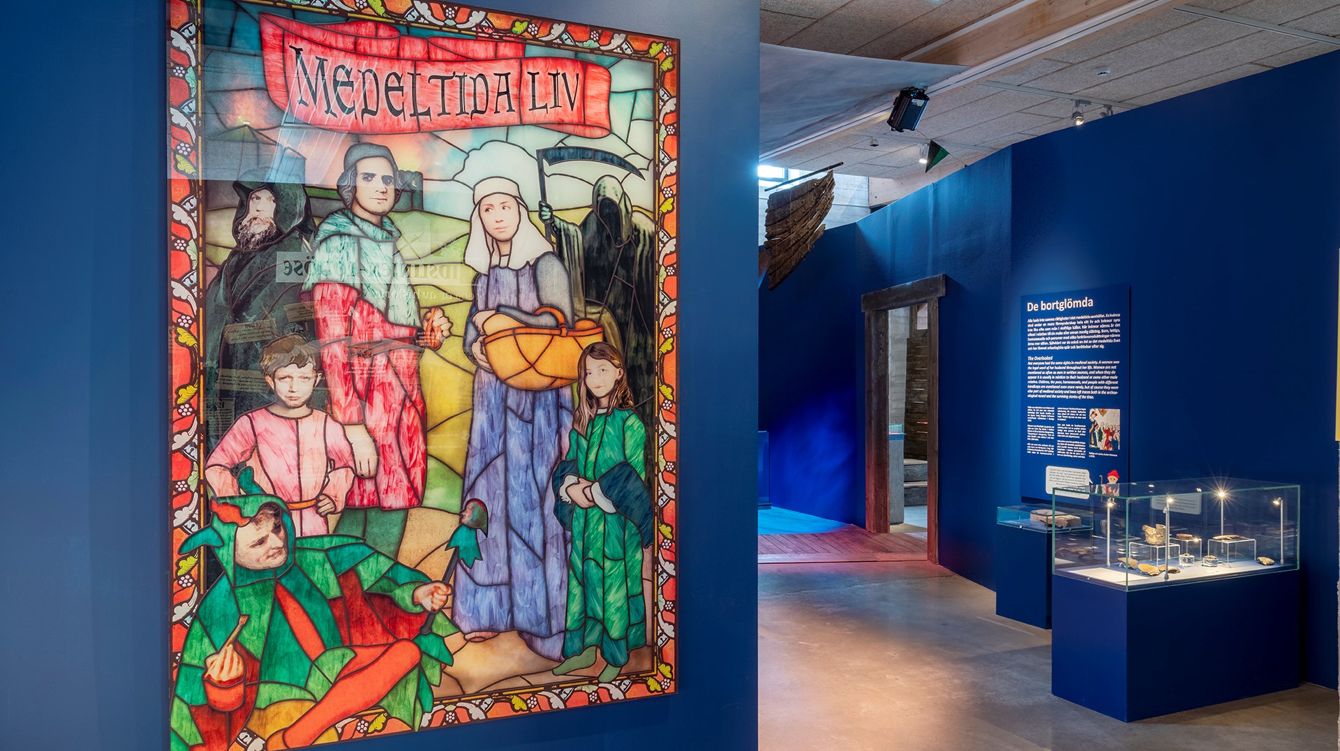 Ett blått rum, med en målad bil i förgrunden. På bilden syns människor i medeltidskläder. I bakgrunden ser man en monter på håll, med föremål i.
