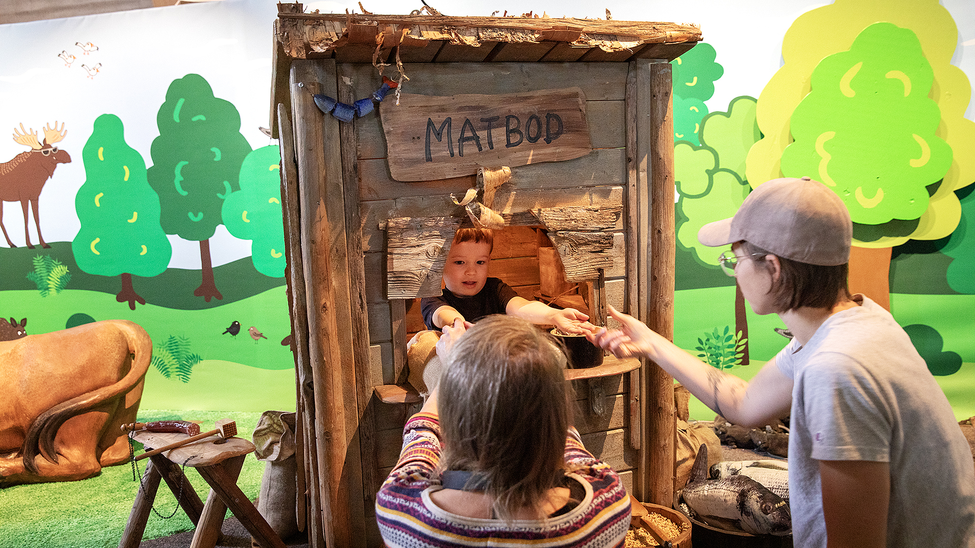 Ett barn står i en kioskliknande bod och räcker två vuxna utanför någonting, kanske leker de affär