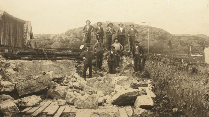 An old picture of men posing in front of a pile of stone and human skulls.