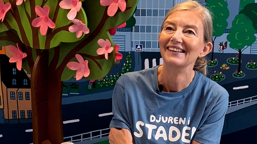Författaren Sarah Sheppard ler mot kameran. I bakgrunden ses en färgglad fond som föreställer träd i en stad.