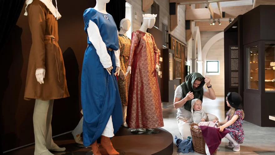 Det är ett stort rum med bruna väggar. I förgrunden syns skyltdockor med medeltida kläder. Bredvid dem sitter en pappa med sina två små barn och provar medeltida hattar och mössor.