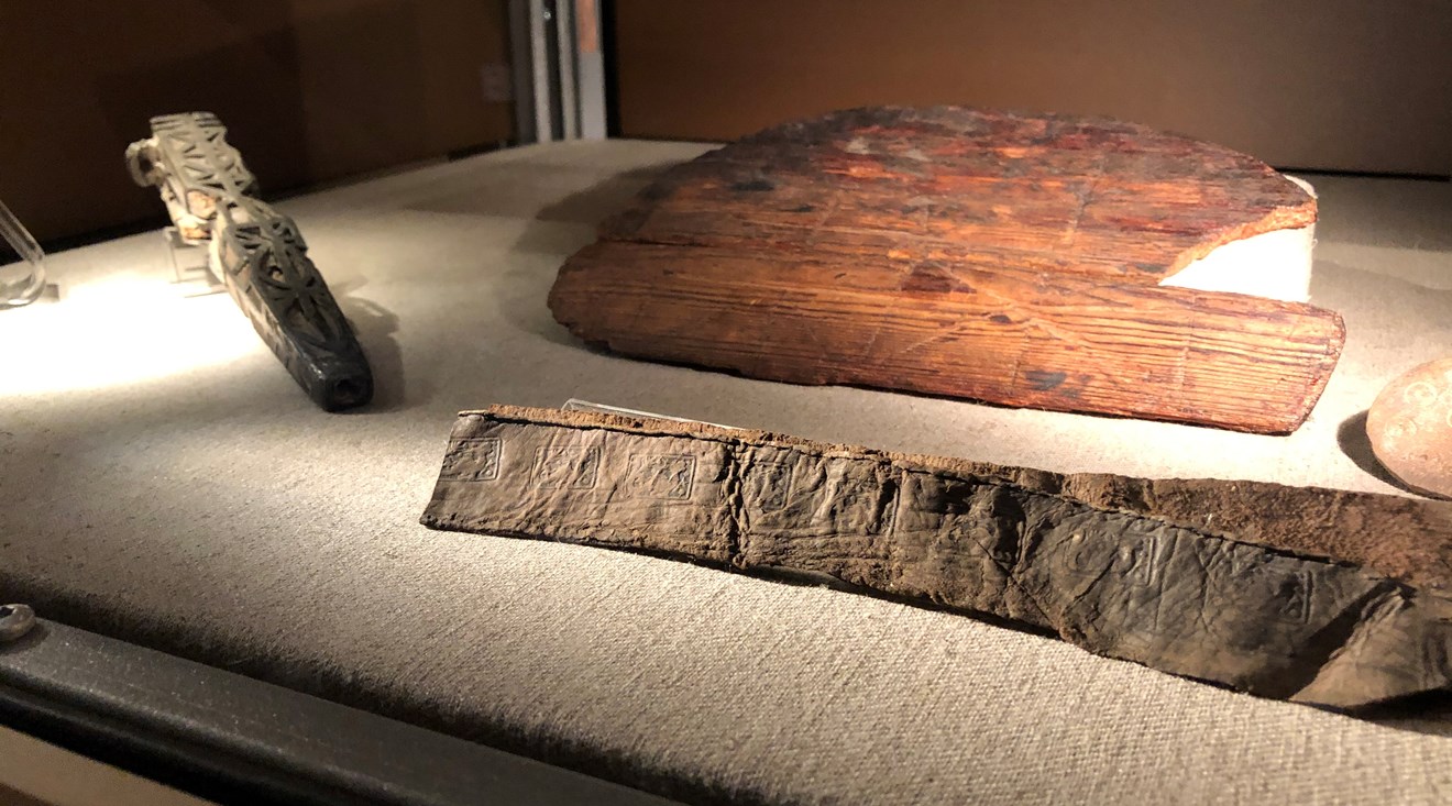 En närbild på medeltida föremål i en monter. Det är ett knivfodral i trä, en svärdsskida i läder, och ett spelbräde i trä.