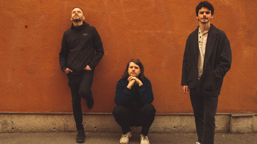 Artisterna i Jonatan Stirner trio står lutade mot en orange vägg