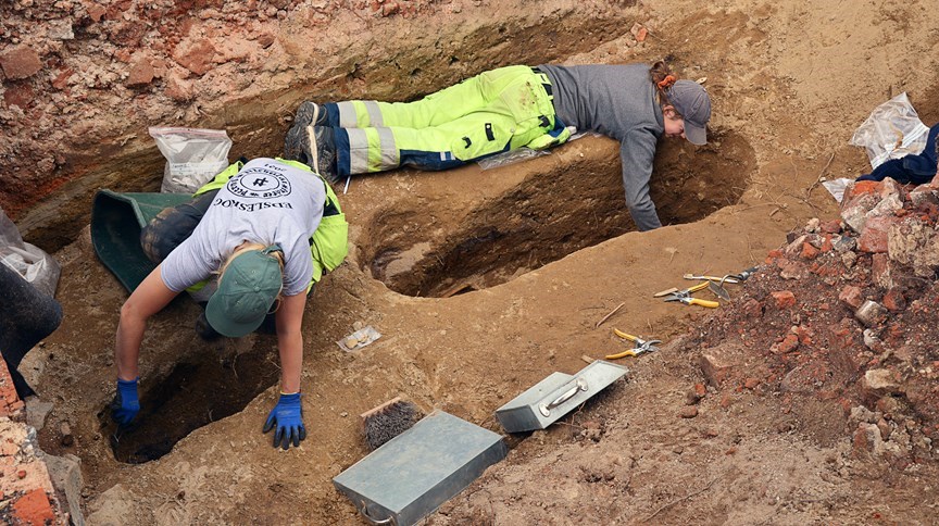 Två arkeologer ligger på marken och gräver i var sin grop