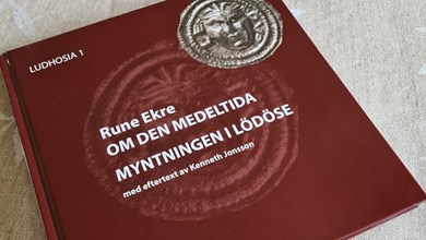 En röd bok med titel "Om den medeltida myntningen i Lödöse" ligger på en ljus duk på ett bord