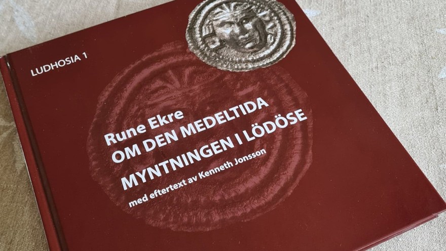 En röd bok med titel "Om den medeltida myntningen i Lödöse" ligger på en ljus duk på ett bord