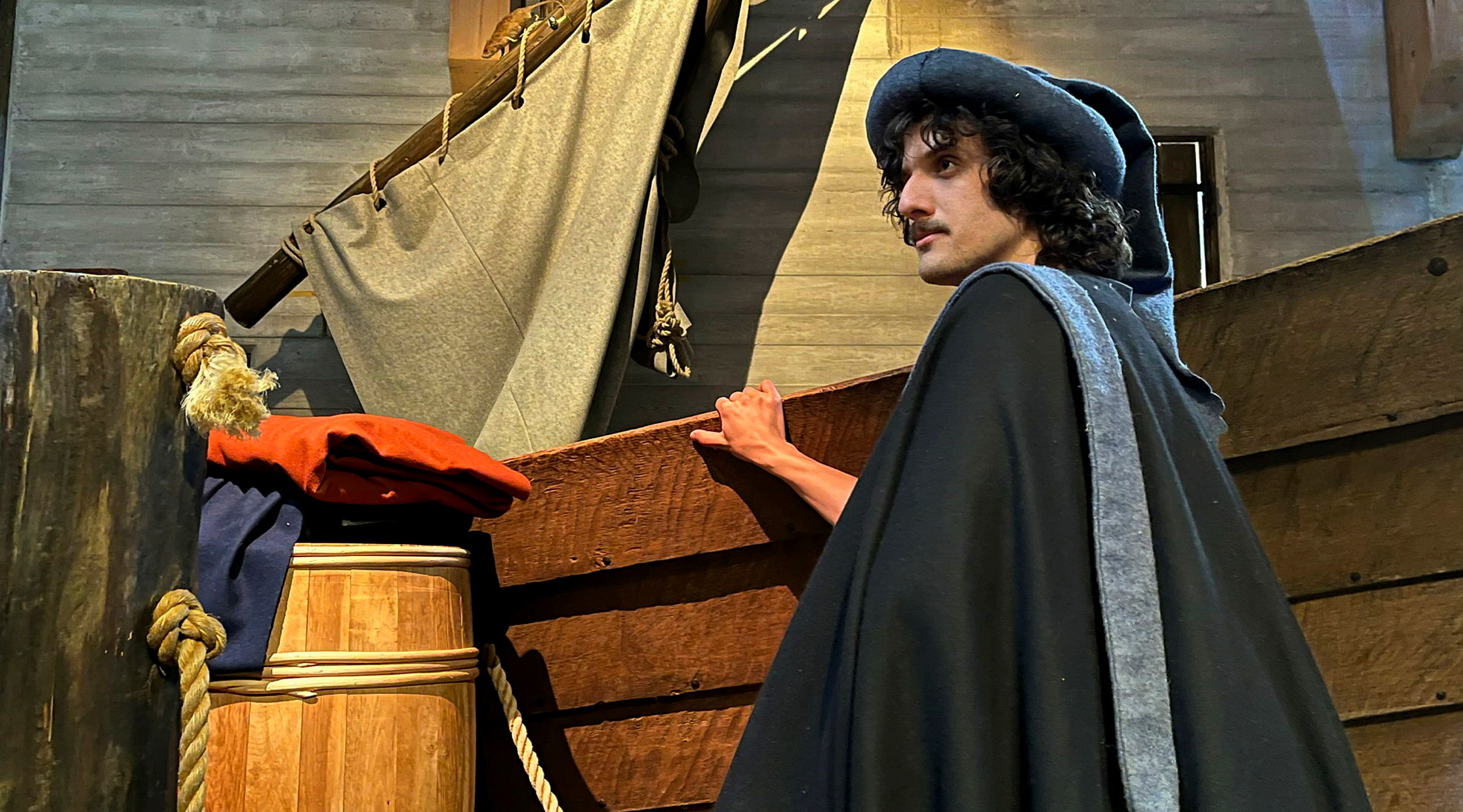 En man står vänd från kameran. Han står mer till höger i bild och har en svart cape på sig. Vi ser hans ansikte i profil. Han har mörkt hår och en huvudbonad på huvudet. Han håller i en träpåle, en uppbyggd del av en hamn. Vi ser en stor trätunna och en liten del av en segelbåt.