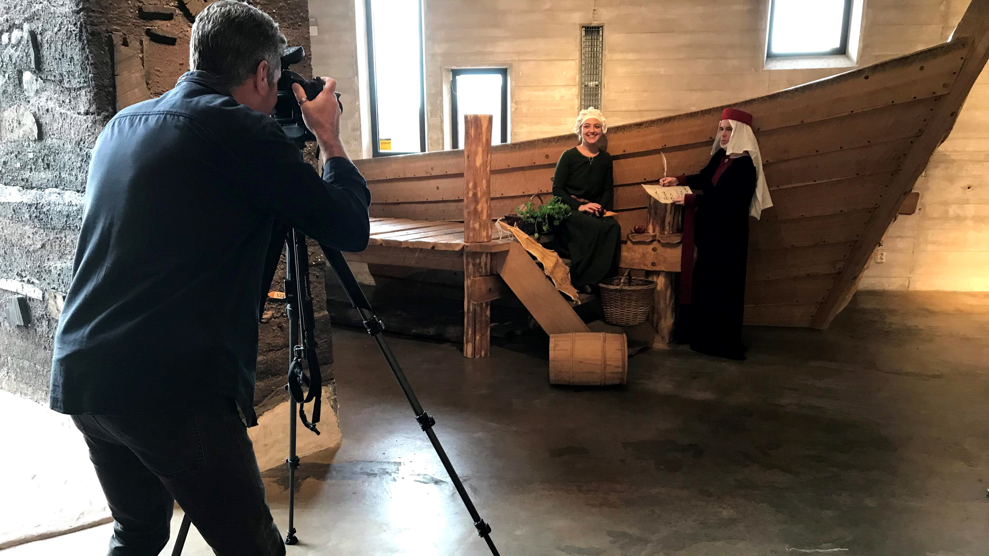 En man fotar två kvinnor som är klädda i medeltidskläder. Kvinnorna står och sitter framför en båt inne i en utställningshall.