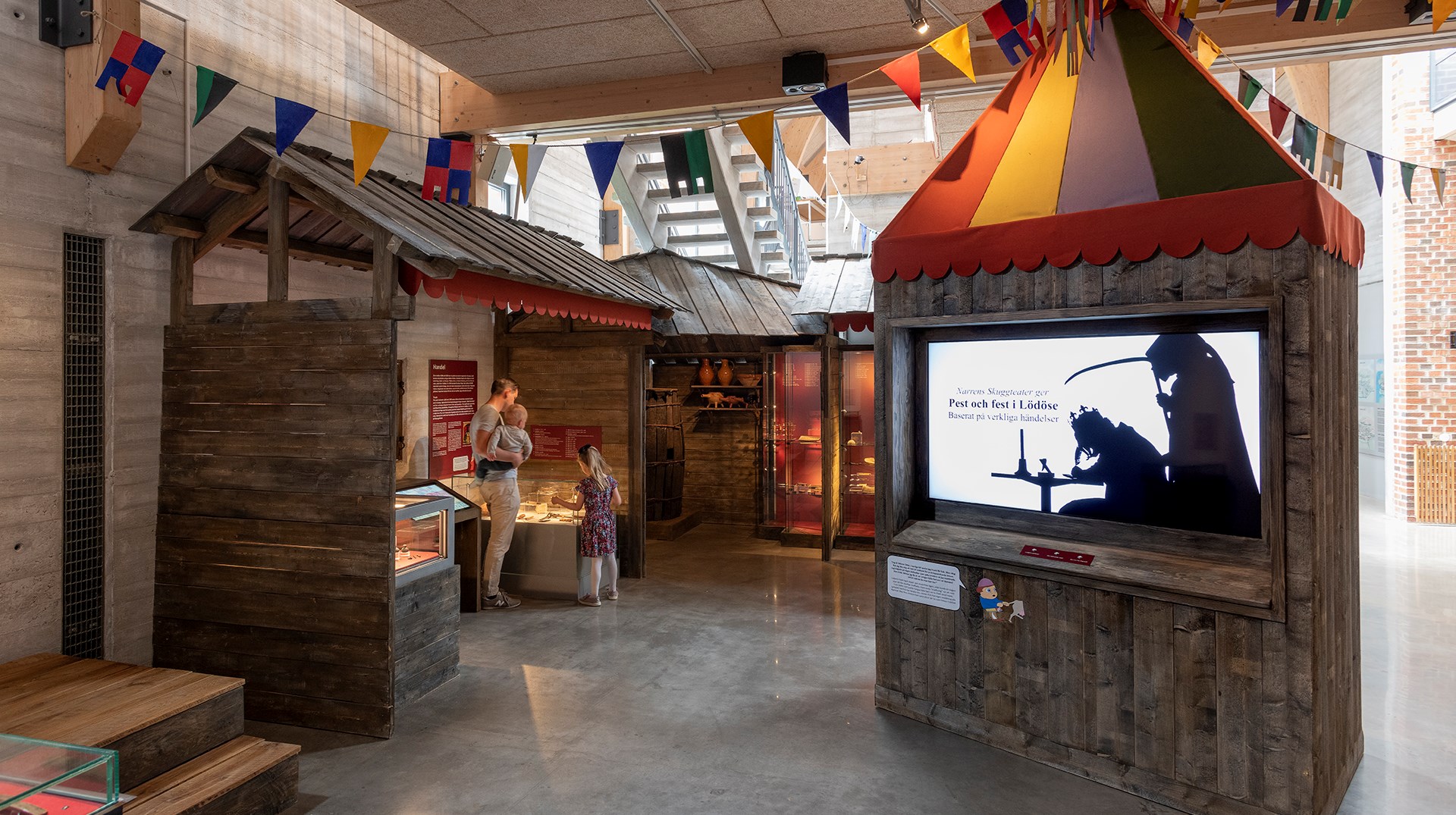 Det är en översiktsbild över ett rum som kallas "Torget". Det är uppbyggda miljöer som ska föreställa medeltida handelsbodar. I bodarna finns montrar med föemål. I mitten av rummet står en konstruktion av trä med en in byggd skärm. På skärmen visas filmer i form av skuggspel.