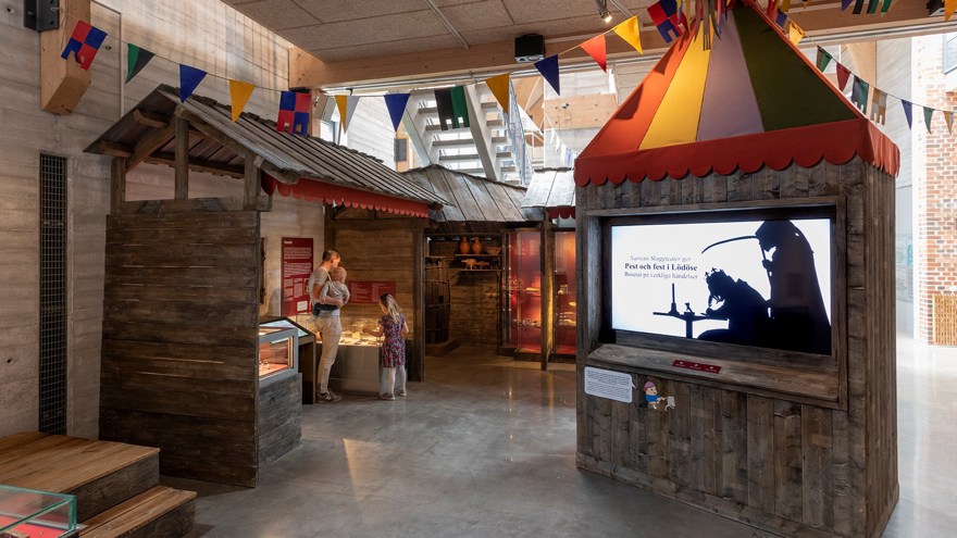 Det är en översiktsbild över ett rum som kallas "Torget". Det är uppbyggda miljöer som ska föreställa medeltida handelsbodar. I bodarna finns montrar med föemål. I mitten av rummet står en konstruktion av trä med en in byggd skärm. På skärmen visas filmer i form av skuggspel.