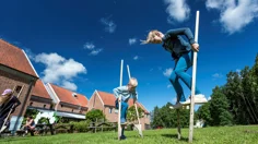 Två barn går på styltor på en gräsmatta en solig dag. I bakgrunden syns en röd tegelbyggnad