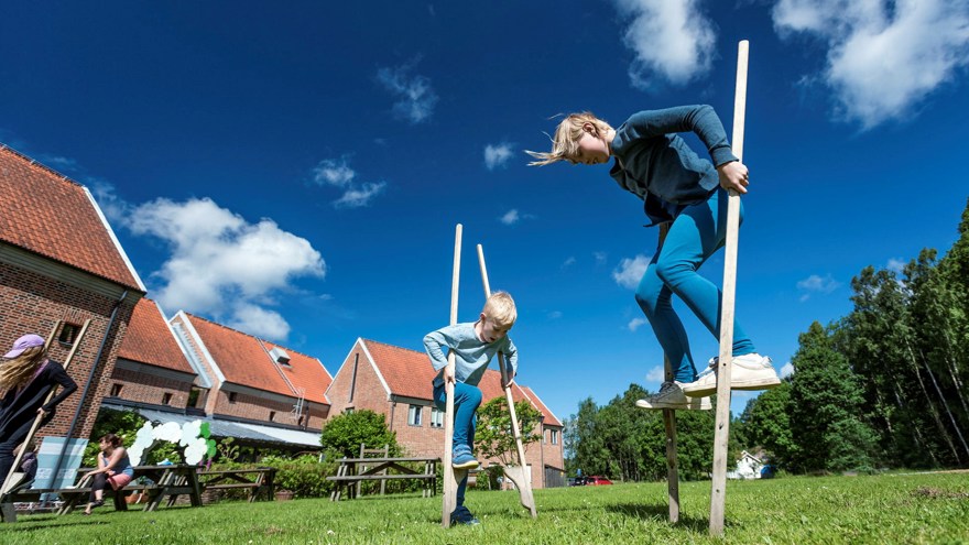 Två barn går på styltor på en gräsmatta en solig dag. I bakgrunden syns en röd tegelbyggnad