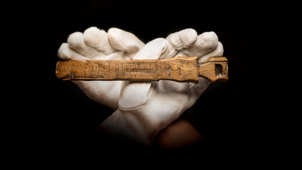 Svart bakgrund, två korsade händer i vita handskar håller en snidad skälkniv. 